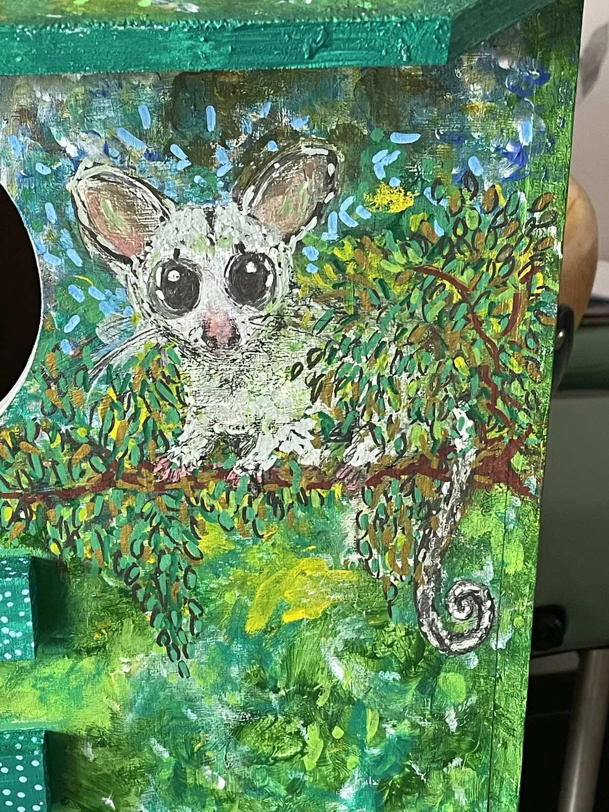 Close-up of the painted possum with big dark eyes and a curled tail, sitting among green leaves
