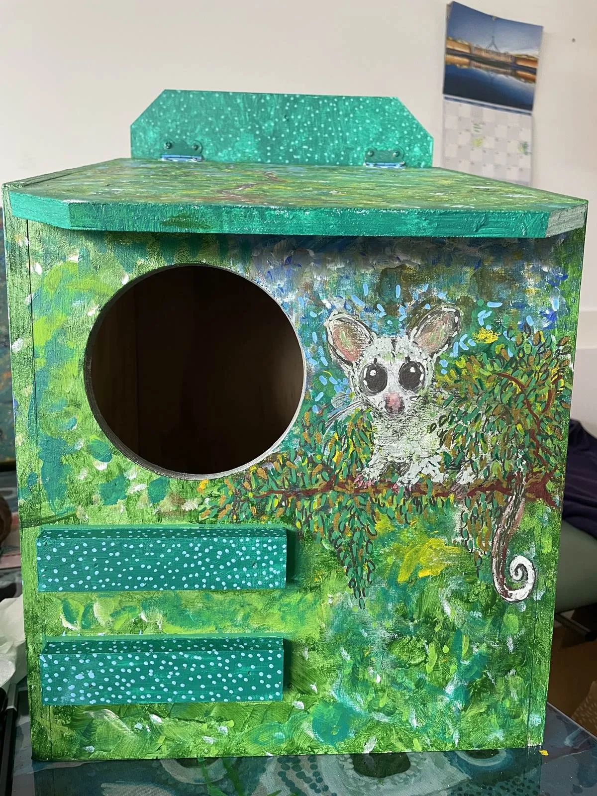 Front of a hand-painted possum nest box: a round entry hole beside a painted possum on a leafy branch, in bright greens
