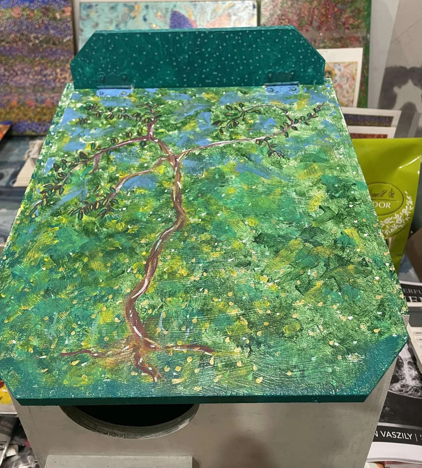 Hinged lid of the possum box painted with a brown tree and green canopy against a blue sky